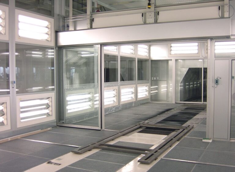 Cleanroom-style industrial spray booth with glass partitions and overhead lighting.